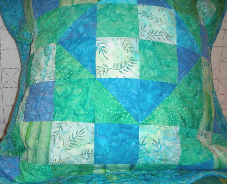 Puede incluir: Una funda de coj&iacute;n acolchada cuadrada con un dise&ntilde;o de patchwork de varios cuadrados de tela azul y verde. Los patrones de la tela incluyen colores s&oacute;lidos, motivos de hojas y texturas punteadas, unidos por costuras blancas visibles en un patr&oacute;n ondulado.