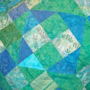 Puede incluir: Una colcha de patchwork con un patr&oacute;n geom&eacute;trico de tri&aacute;ngulos y cuadrados en tonos azules, verde azulado y verdes. La tela muestra varios estampados estilo batik con motivos de hojas y puntos, con acolchado a m&aacute;quina en l&iacute;neas onduladas.