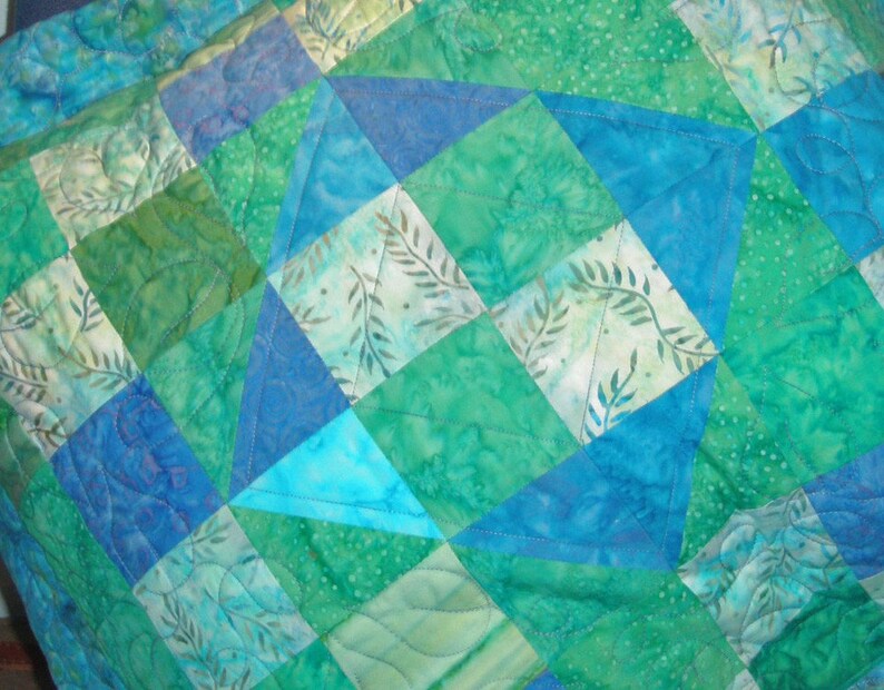 Puede incluir: Una colcha de patchwork con un patr&oacute;n geom&eacute;trico de tri&aacute;ngulos y cuadrados en tonos azules, verde azulado y verdes. La tela muestra varios estampados estilo batik con motivos de hojas y puntos, con acolchado a m&aacute;quina en l&iacute;neas onduladas.