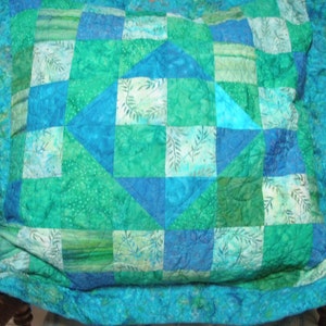 Puede incluir: Una funda de coj&iacute;n acolchada cuadrada con un dise&ntilde;o de patchwork en varios tonos de azul y verde. La tela muestra una mezcla de colores s&oacute;lidos y estampados de hojas, con un motivo central de diamante rodeado por bloques cuadrados m&aacute;s peque&ntilde;os.
