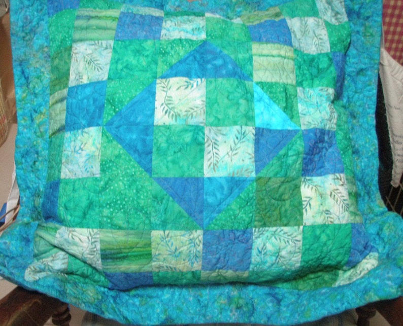 Puede incluir: Una funda de coj&iacute;n acolchada cuadrada con un dise&ntilde;o de patchwork en varios tonos de azul y verde. La tela muestra una mezcla de colores s&oacute;lidos y estampados de hojas, con un motivo central de diamante rodeado por bloques cuadrados m&aacute;s peque&ntilde;os.