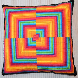 May include: A colorful pillow with a geometric pattern of squares and stripes in shades of red, orange, yellow, green, blue, and purple. The pillow is made of fabric and has a black border.