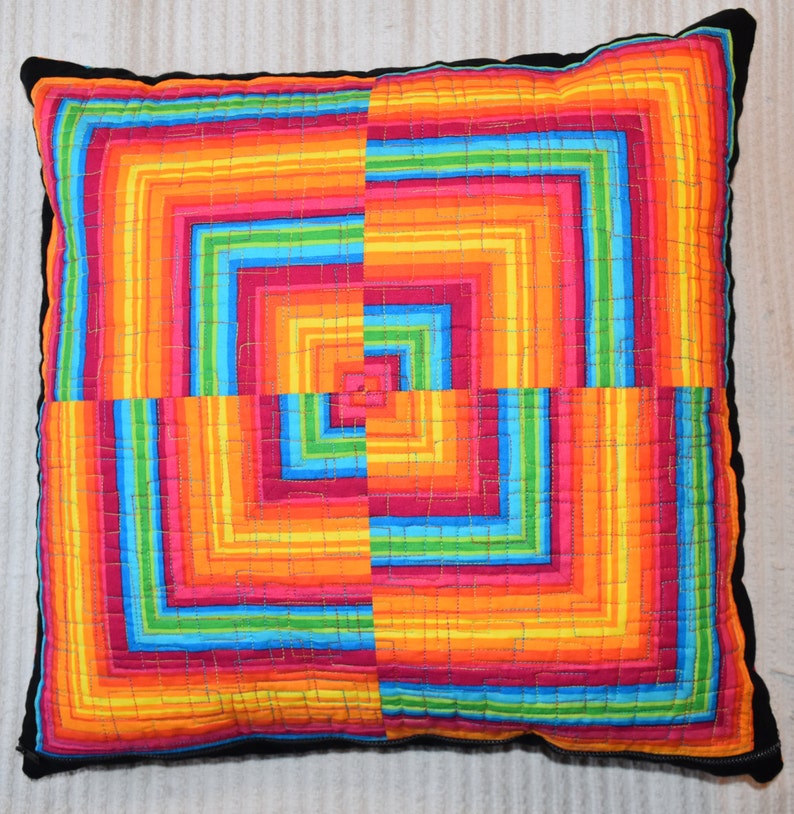 May include: A colorful pillow with a geometric pattern of squares and stripes in shades of red, orange, yellow, green, blue, and purple. The pillow is made of fabric and has a black border.