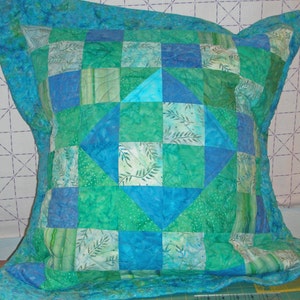 Puede incluir: Una funda de coj&iacute;n acolchada cuadrada con un dise&ntilde;o de patchwork en tonos azul verdoso, turquesa y verde esmeralda. La tela muestra varios patrones que incluyen hojas y puntos, con un motivo de diamante central y un borde ancho acolchado.