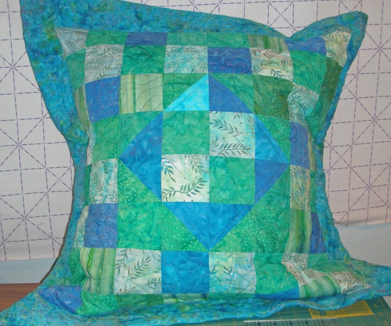 Puede incluir: Una funda de coj&iacute;n acolchada cuadrada con un dise&ntilde;o de patchwork en tonos azul verdoso, turquesa y verde esmeralda. La tela muestra varios patrones que incluyen hojas y puntos, con un motivo de diamante central y un borde ancho acolchado.