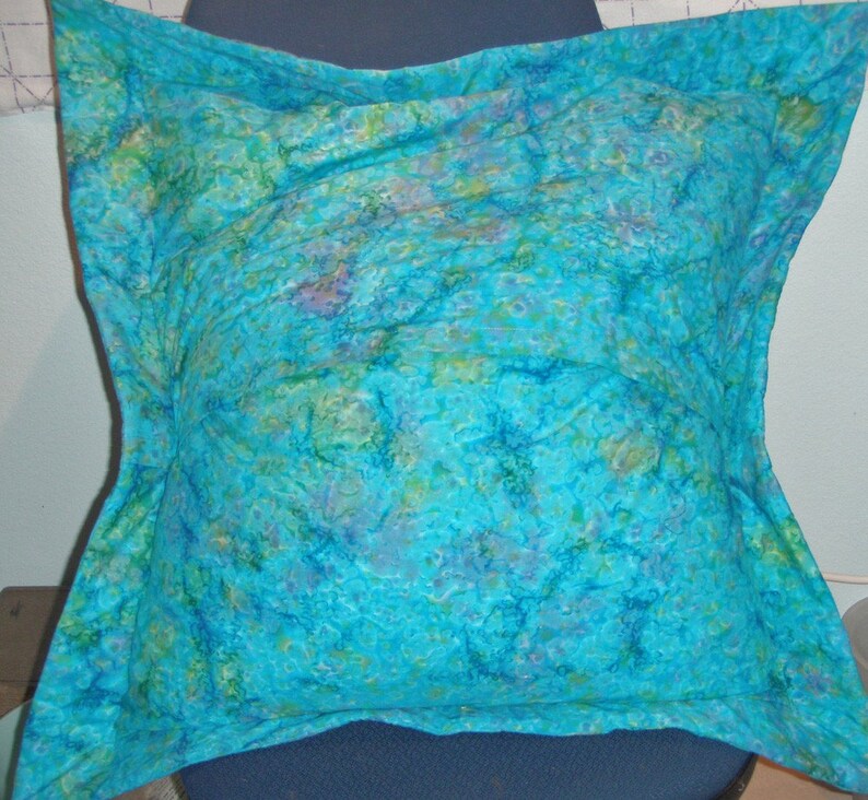 Puede incluir: Un coj&iacute;n cuadrado con un vibrante patr&oacute;n jaspeado en tonos turquesa, azul y verde. La tela presenta una apariencia texturizada y moteada con suaves transiciones de color en toda la superficie. El coj&iacute;n tiene un dise&ntilde;o de borde distintivo.
