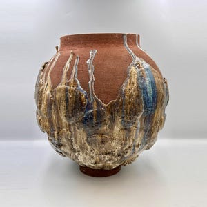 Op de afbeelding: Een ronde keramische vaas met een brede opening en een mat terracotta-kleurig bovengedeelte. Het onderste gedeelte heeft een dik, getextureerd glazuur in tinten crème, bruin en blauw dat vanuit het midden naar de basis toe naar beneden druipt.