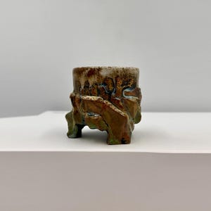 Op de afbeelding: Een kleine, cilindervormige keramische plantenpot met een ruwe, onregelmatige textuur. Het oppervlak heeft een gevlekt glazuur in aardse tinten bruin, beige en accenten van blauwgroen. De basis rust op drie opvallende, dikke, onregelmatige voetjes.
