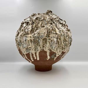 Può includere: Una scultura sferica in ceramica con una superficie ruvida e strutturata. La parte inferiore è in argilla bruno-rossastra, mentre la sezione superiore presenta uno spesso smalto bianco che cola su una texture rocciosa, marrone e grigia.