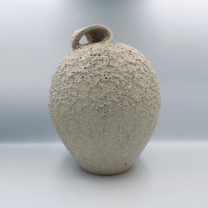 Può includere: Un vaso in ceramica color bianco sporco di forma rotonda con una superficie fortemente strutturata e craterizzata che ricorda un paesaggio lunare. Il vaso presenta un unico piccolo manico arcuato sulla parte superiore, integrato nel corpo rotondo.