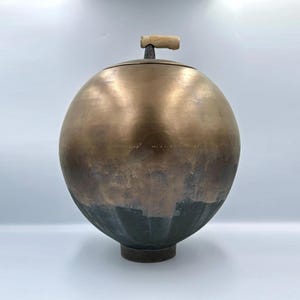 Könnte beinhalten: Ein kugelförmiges Gefäß mit einem metallischen Bronzefinish auf der oberen Hälfte und einem dunklen, texturierten anthrazitfarbenen Finish auf der unteren Hälfte. Das Objekt hat einen kleinen, hellen zylindrischen Griff oben und steht auf einem runden Sockel.