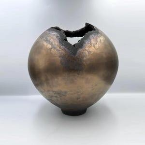 May include: A spherical bronze-toned vessel with a smooth, metallic finish and a dark, jagged, uneven opening at the top. The surface features mottled brown and copper-colored textures, tapering down to a small, circular base at the bottom.