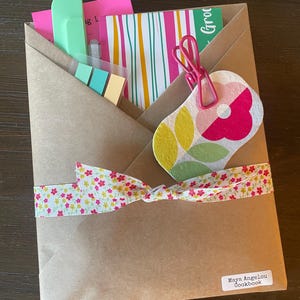 May include: A brown paper envelope tied with a floral ribbon, containing a pink card, a striped card, a mint green tool, and sticky notes. A pink clip holds a flower-shaped tag with green and pink petals. A label reads Maya Angelou Cookbook.