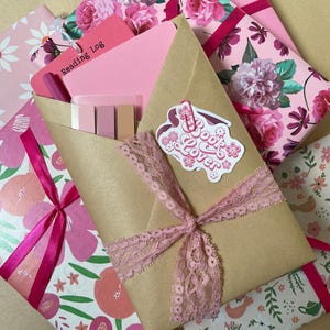 May include: An assortment of pink and floral-patterned stationery items. A brown envelope tied with pink lace and a ribbon features a "Book Lover" sticker. A pink tag reads "Reading Log".