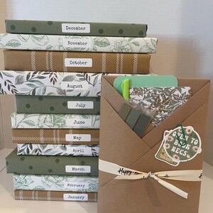 May include: A stack of twelve books wrapped in patterned paper, each labeled with a month from January to December. Beside the stack is a brown paper-wrapped package tied with a ribbon, containing a green pen, sticky tabs, and a sticker.