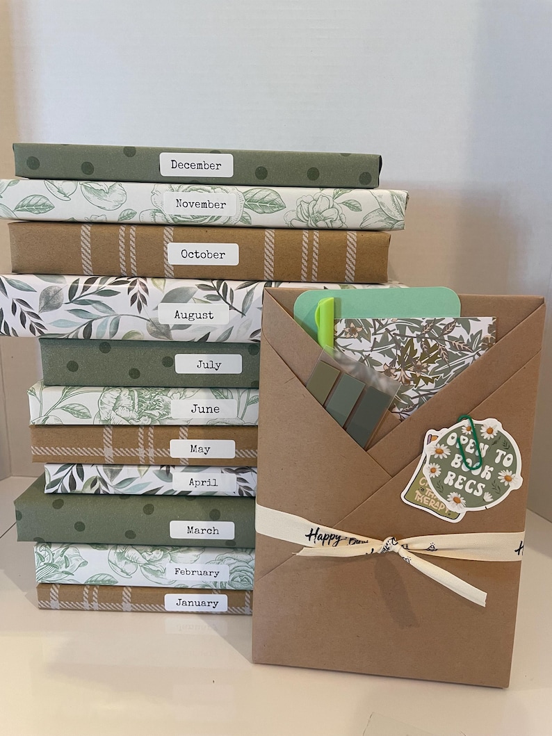 May include: A stack of twelve books wrapped in patterned paper, each labeled with a month from January to December. Beside the stack is a brown paper-wrapped package tied with a ribbon, containing a green pen, sticky tabs, and a sticker.