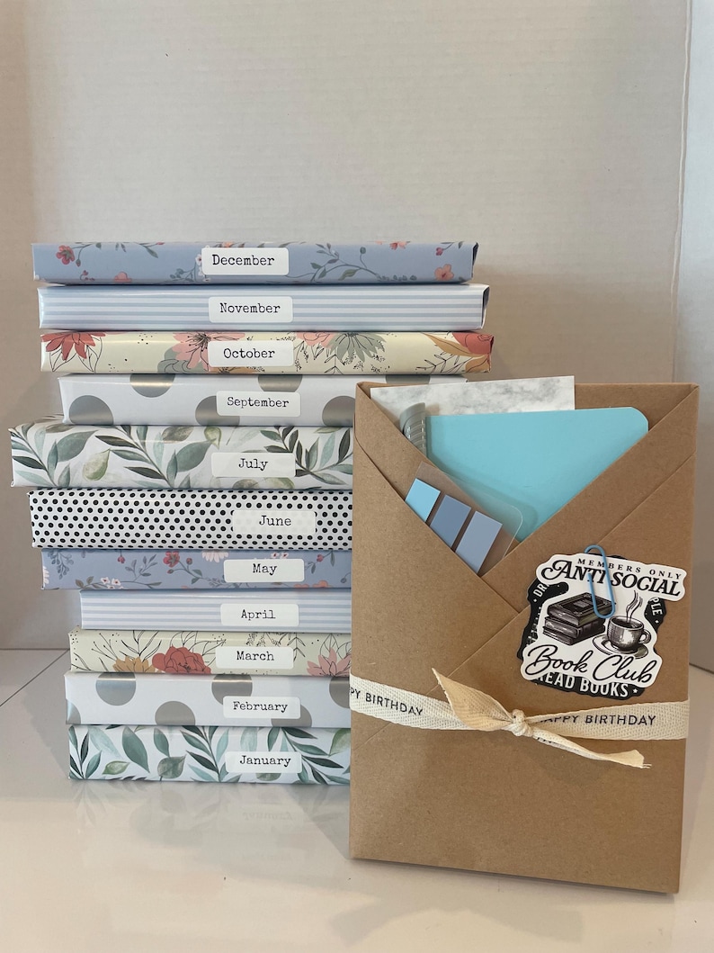 May include: A stack of twelve books wrapped in patterned paper, each labeled with a month from January to December. Beside the stack is a brown paper envelope containing stationery, featuring a sticker that reads Anti Social Book Club.