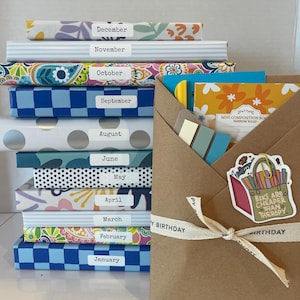 May include: A stack of twelve notebooks with patterned covers, each labeled with a month from January to December. Beside the stack is a brown paper envelope tied with a ribbon, containing a mini composition book and a sticker reading Books Are Cheaper Than Therapy.