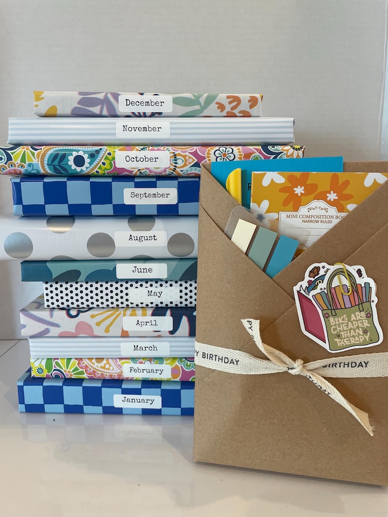 May include: A stack of twelve notebooks with patterned covers, each labeled with a month from January to December. Beside the stack is a brown paper envelope tied with a ribbon, containing a mini composition book and a sticker reading Books Are Cheaper Than Therapy.