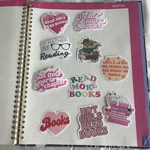May include: A spiral-bound notebook open to a page with several colorful stickers. The stickers feature phrases like "Good Girls Read Books," "Read More Books," and "I'd Rather Be Reading." The stickers are in various shapes and colors.