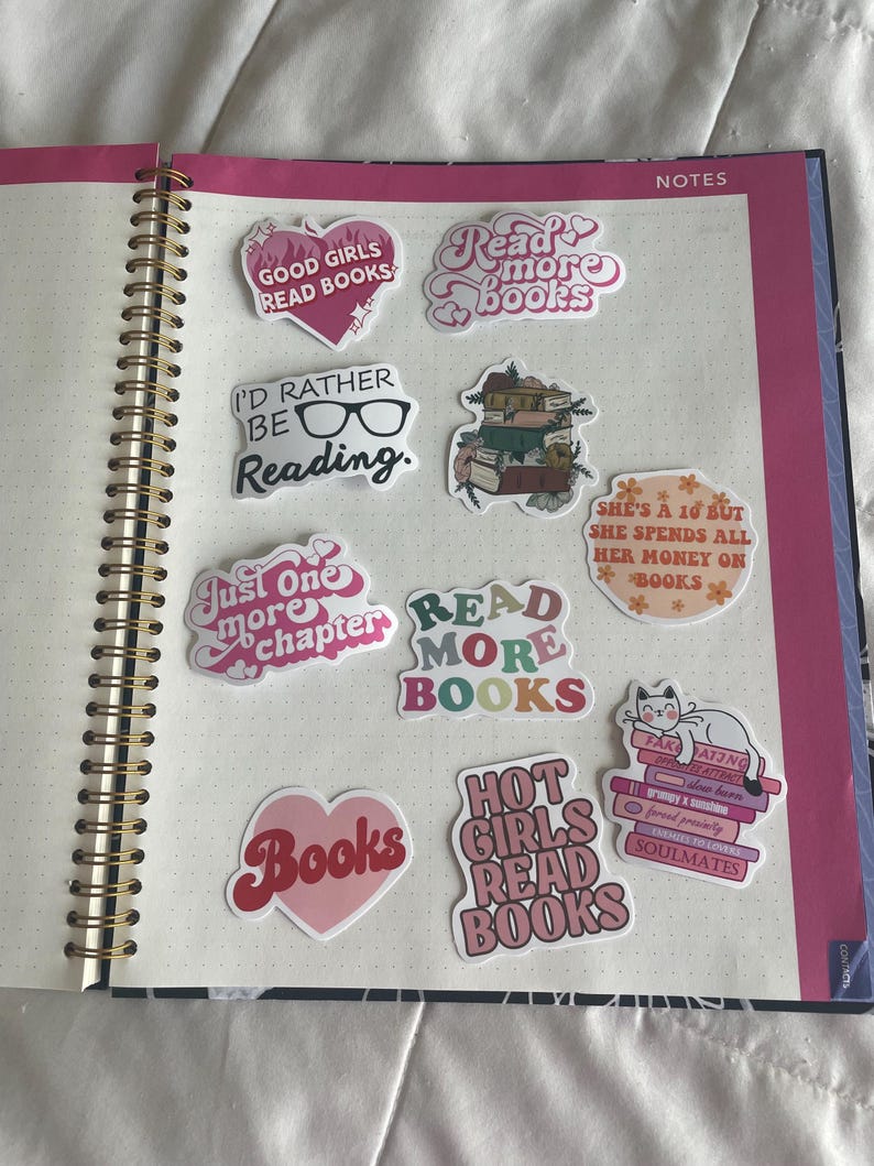 May include: A spiral-bound notebook open to a page with several colorful stickers. The stickers feature phrases like "Good Girls Read Books," "Read More Books," and "I'd Rather Be Reading." The stickers are in various shapes and colors.