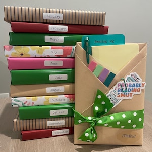 May include: A stack of twelve brown, red, green, and pink wrapped books labeled with the months of the year. A brown paper package with a green ribbon and white polka dots is tied with a bow. The package is labeled "Reading Log" and "Romance". A sticker with the text "Probably Reading Smut" is on the package.