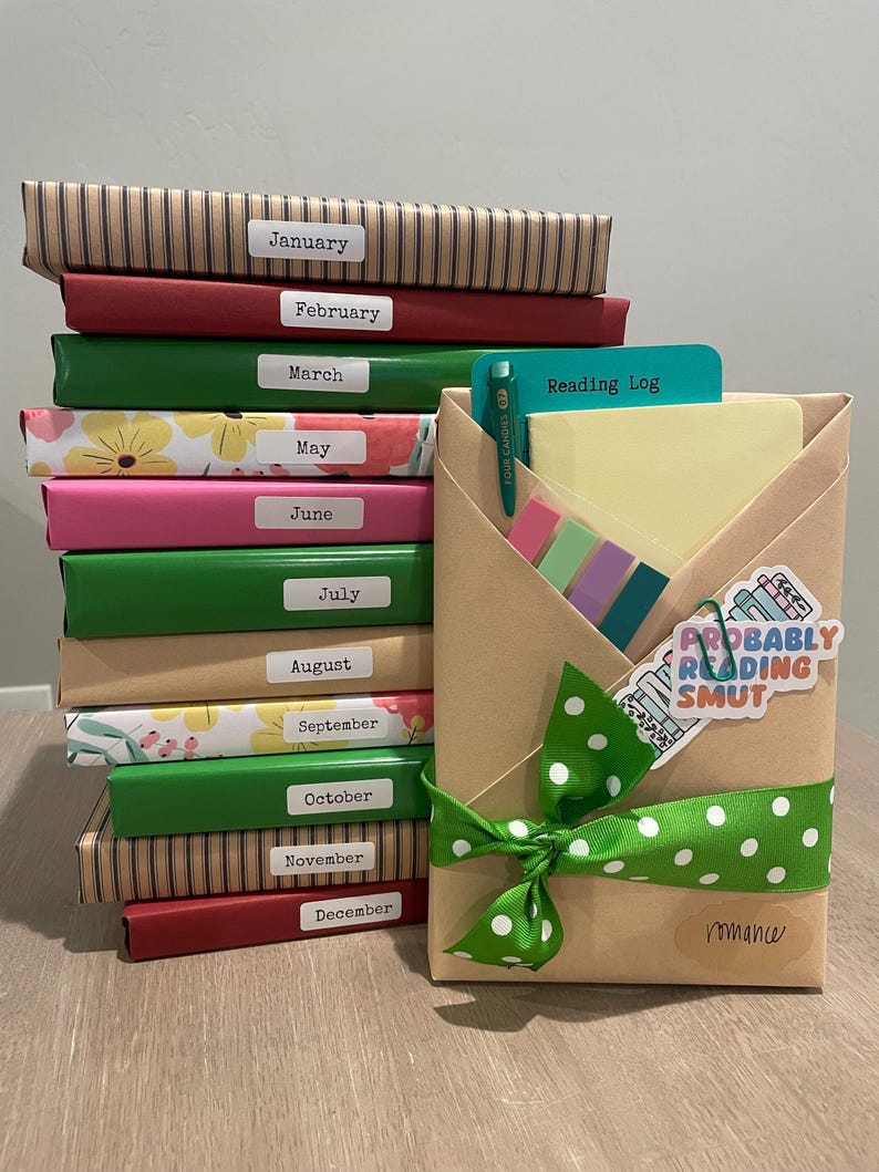 May include: A stack of twelve brown, red, green, and pink wrapped books labeled with the months of the year. A brown paper package with a green ribbon and white polka dots is tied with a bow. The package is labeled "Reading Log" and "Romance". A sticker with the text "Probably Reading Smut" is on the package.
