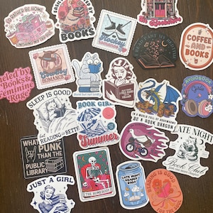 May include: A collection of colorful stickers with various designs and text. Themes include books, reading, coffee, and book clubs. The stickers feature illustrations of cats, skeletons, and other whimsical elements. The stickers are on a dark brown surface.