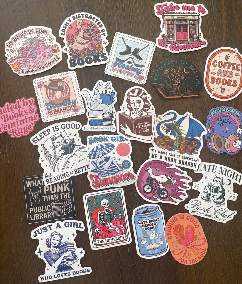 May include: A collection of colorful stickers with various designs and text. Themes include books, reading, coffee, and book clubs. The stickers feature illustrations of cats, skeletons, and other whimsical elements. The stickers are on a dark brown surface.