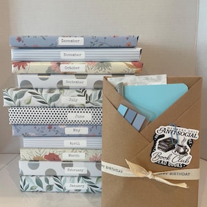 May include: A stack of twelve books wrapped in patterned paper, each labeled with a month from January to December. Beside the stack is a brown paper envelope containing stationery, featuring a sticker that reads Anti Social Book Club.