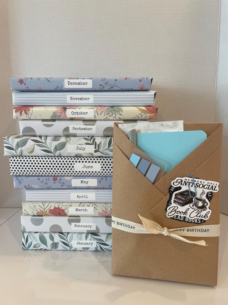 May include: A stack of twelve books wrapped in patterned paper, each labeled with a month from January to December. Beside the stack is a brown paper envelope containing stationery, featuring a sticker that reads Anti Social Book Club.