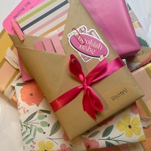 May include: A stack of wrapped gifts with floral and striped paper. The top gift is wrapped in brown paper with a pink ribbon and a sticker that says "bookish babe".