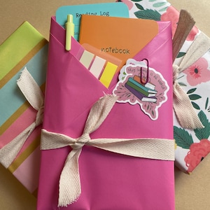 May include: A bright pink paper-wrapped package tied with a cream-colored ribbon, containing an orange notebook labeled "notebook," a teal "Reading Log" card, a yellow pen, and sticky tabs. A sticker with books and the text "My summer is all booked" is attached.
