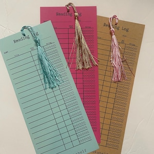 May include: Three colorful reading log bookmarks. The bookmarks are light blue, pink, and brown. Each bookmark has a tassel and the words "Reading Log" printed at the top. The bookmarks have lines for the reader to fill in the title, start date, and rating.