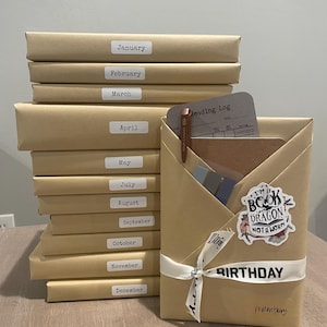 May include: A stack of twelve brown wrapped books with labels for each month of the year. The top book is wrapped in brown paper with a white ribbon and a sticker that says "I'm a book dragon, not a worm". The book is labeled "Birthday" and "romantasy".