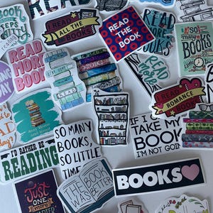 May include: A collection of colorful stickers with book-related themes. The stickers feature phrases like "Read," "Books," and "Just one more chapter." Some stickers have illustrations of books, cats, and bookshelves. The stickers are in various shapes and sizes.