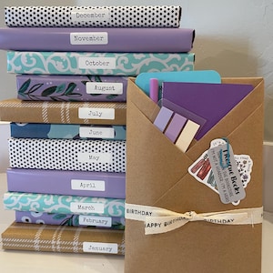 May include: A stack of twelve books wrapped in various patterned papers, each labeled with a month from January to December. Beside the stack is a brown paper envelope containing a purple notebook, sticky notes, and a "Rescue Books" tag.