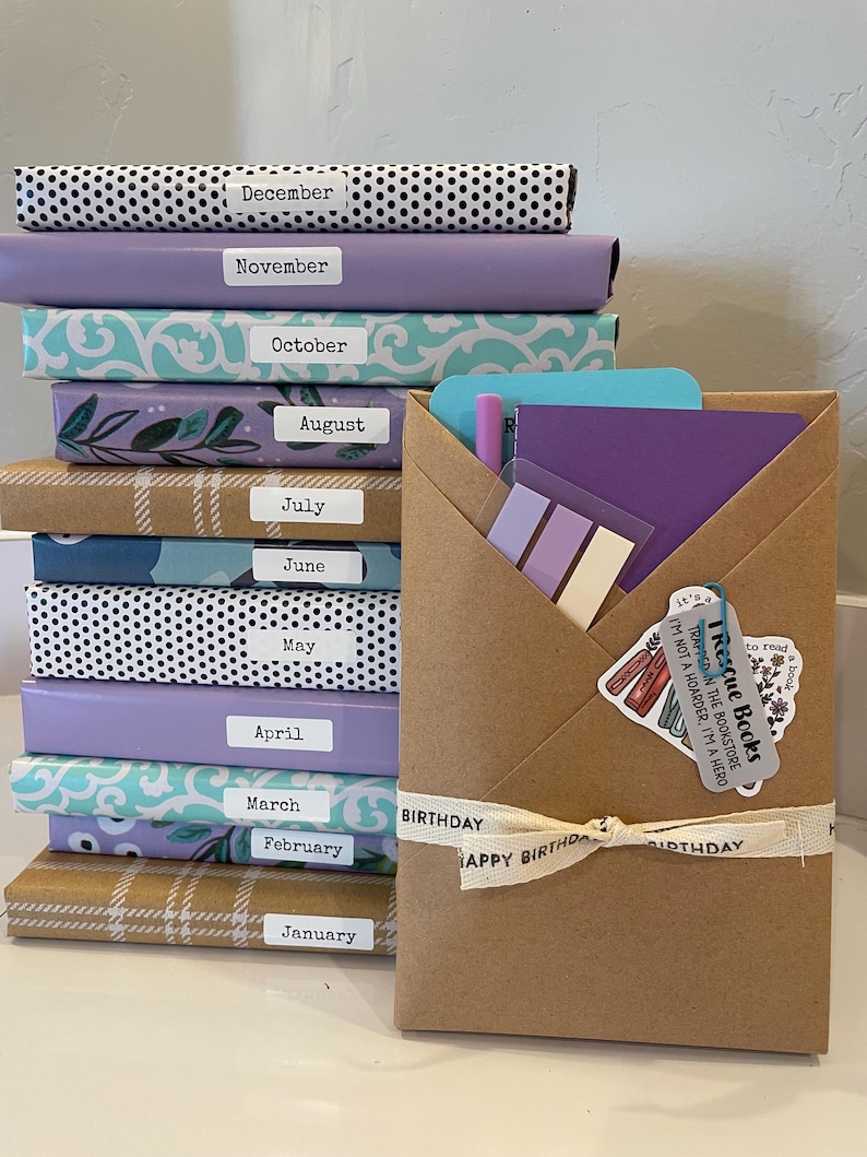 May include: A stack of twelve books wrapped in various patterned papers, each labeled with a month from January to December. Beside the stack is a brown paper envelope containing a purple notebook, sticky notes, and a "Rescue Books" tag.