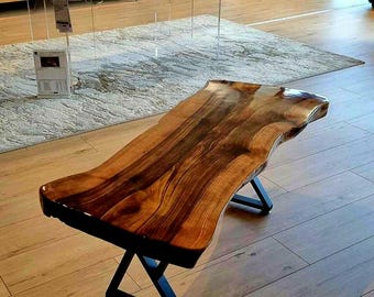 Puede incluir: Una mesa de centro con borde vivo y acabado de vetas de madera oscura. La mesa está sostenida por patas de metal negro con diseño en forma de X. La parte superior de la mesa tiene una forma natural y orgánica.