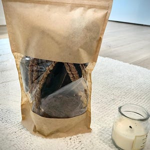 Peut inclure: Un sac en papier brun avec une fenêtre transparente contenant des articles brun foncé est posé sur une surface blanche texturée. Une petite bougie en pot de verre avec de la cire blanche et une étiquette est à côté du sac. La bougie a une mèche brûlée.