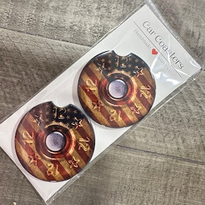May include: Two round car coasters with a red, white, and blue American flag design. The center of each coaster features a brown and gold bullet casing with the number "12" printed on it. The text "Car Coasters" and "Handmade with love" is printed on a white card behind the coasters.