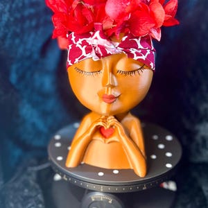 May include: A decorative planter shaped like a woman's head, with closed eyes and a red heart. The head is adorned with a red and white patterned headband and topped with vibrant red flowers. The planter is on a black base.
