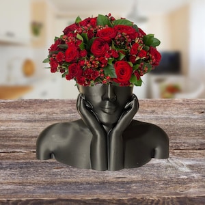 Puede incluir: Un busto decorativo negro con manos sosteniendo la cara, que contiene un ramo de rosas y bayas rojas. Este jarrón único es perfecto para arreglos florales. Las flores rojas vibrantes contrastan con la oscura escultura.