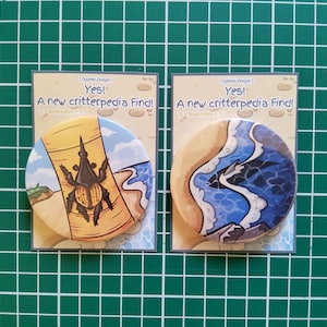 May include: Two round buttons with a white background and colourful illustrations. The first button features a brown and yellow beetle on a brown and yellow tree trunk. The second button features a blue and white wave with a black and white creature inside. Both buttons have the text "Yes! A new critterpedia Find!" on the white background.