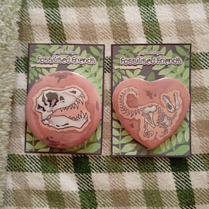 May include: Two pin-back badges on display cards labelled Fossilised Friends. One badge is circular with a T-Rex skull illustration, and the other is heart-shaped with a full T-Rex skeleton illustration. Both feature a dusty pink background.