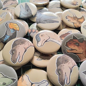 May include: A pile of circular buttons, each with a dinosaur design. The illustrations are in shades of brown, grey, and blue, set against beige and grey backgrounds. The buttons are a mix of different dinosaur species.