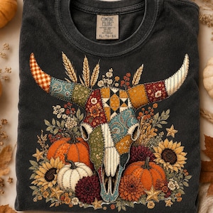 Peut inclure: Un t-shirt gris anthracite orné d'un motif central représentant un crâne de bœuf décoré d'un patchwork de tissus aux tons automnaux. Le crâne est entouré de citrouilles, de tournesols, d'épis de blé et d'étoiles dans des tons orange et brun.