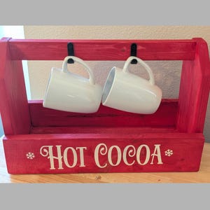 May include: A red wooden hot cocoa mug holder with two white ceramic mugs hanging from black metal hooks. The holder has the words "HOT COCOA" in white script.
