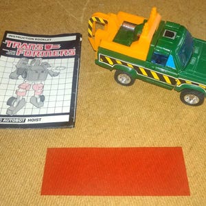 Könnte beinhalten: Grüner und orangefarbener Spielzeug-LKW mit einem schwarz-gelben Streifenmuster. Der LKW hat eine Abschlepphaken und eine Ladefläche. Der LKW ist im Robotermodus. Die Bedienungsanleitung für das Spielzeug ist ebenfalls abgebildet. Die Broschüre enthält eine Schwarzweißillustration des Robotermodus des Spielzeugs. Die Broschüre trägt den Text "INSTRUCTION BOOKLET TRANS FORMERS HEROIC AUTOBOT HOIST".