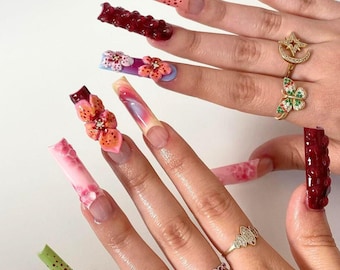 May include: Long, colorful, and decorated acrylic nails with 3D flowers and other designs. The nails are painted in shades of pink, green, red, and white. The nails are decorated with 3D flowers, glitter, and other embellishments.